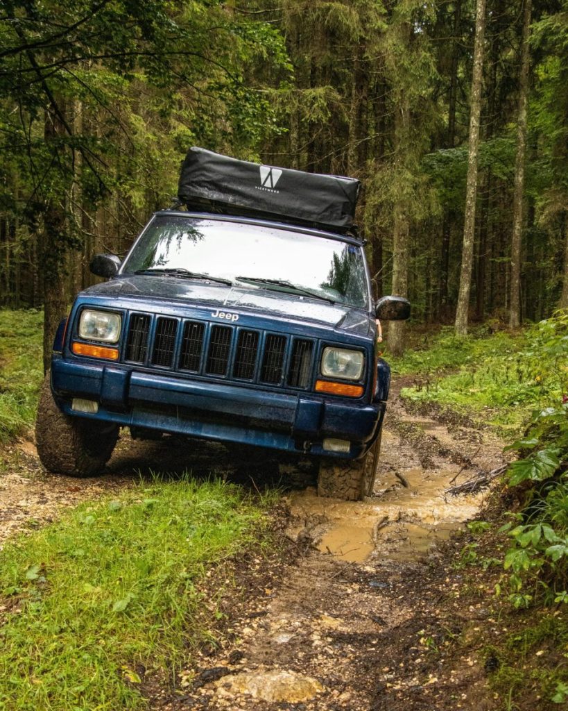 Blue Jeep navigating a muddy off-road trail in the lush forests of Asiago, Italy.